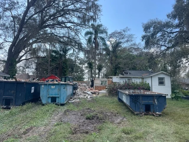 Roll-off dumpsters and rubble pile beside a partially demolished white-sided house with live oaks in background