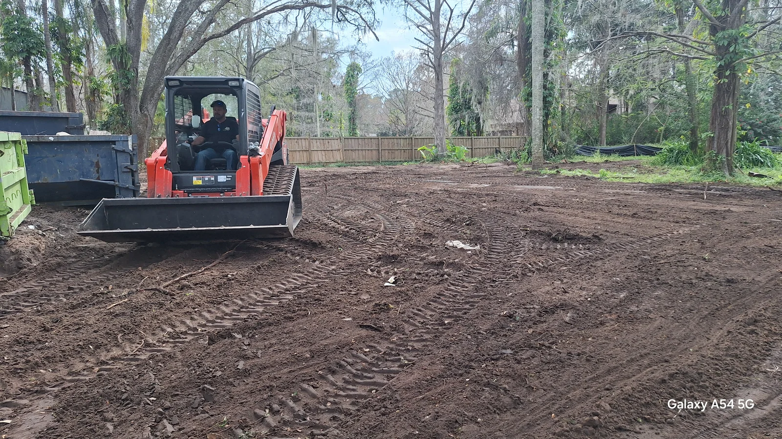 Red compact track loader grading a cleared dirt lot with a flat bucket, with a dumpster and wooden privacy fence in view