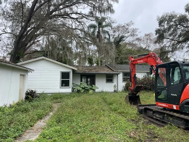 Kubota mini excavator clearing overgrown yard beside white vinyl-sided house with Spanish moss oak trees